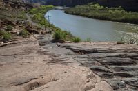 a river flows in the distance next to a cross on a rocky trail as two people walk