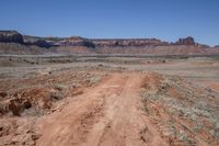 a dirt road through a desert plain with a mountain behind it and a clear blue sky in the background