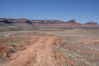 a dirt road through a desert plain with a mountain behind it and a clear blue sky in the background