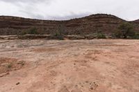 a small field of dirt is near some mountains and grass and bushes with a very steep hill in the background