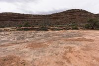 a small field of dirt is near some mountains and grass and bushes with a very steep hill in the background