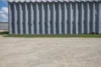 an industrial steel shed next to the grass and the dirt ground and fence with a building