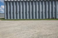 an industrial steel shed next to the grass and the dirt ground and fence with a building