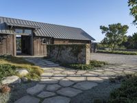 the outside of a rustic house with a walkway through it, a large stone courtyard and two wooden entry doors leading to an open kitchen