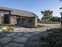 the outside of a rustic house with a walkway through it, a large stone courtyard and two wooden entry doors leading to an open kitchen