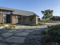 the outside of a rustic house with a walkway through it, a large stone courtyard and two wooden entry doors leading to an open kitchen