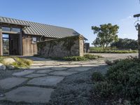 the outside of a rustic house with a walkway through it, a large stone courtyard and two wooden entry doors leading to an open kitchen