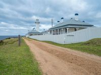 a rural beachside lighthouse in a cloudy sky with light clouds above it and a dirt path going to the building on one side and a gated