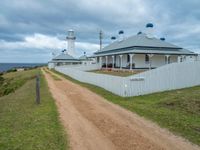 a rural beachside lighthouse in a cloudy sky with light clouds above it and a dirt path going to the building on one side and a gated