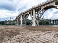 some dirt near a bridge and several buildings, with sky in the background and trees on the far side