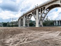 some dirt near a bridge and several buildings, with sky in the background and trees on the far side