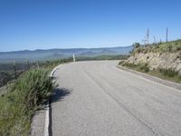 a lone highway on the hillside with a small shrub and mountains in the distance of it