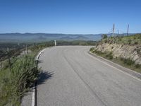 a lone highway on the hillside with a small shrub and mountains in the distance of it