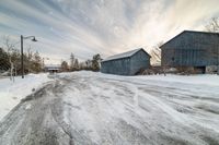 two blue barn sit by the side of a snow covered road and light pole in the distance