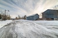 two blue barn sit by the side of a snow covered road and light pole in the distance