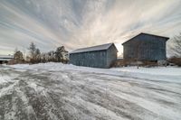 snow is piled near two small barns and one with a sky background is shown at sunset