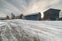 snow is piled near two small barns and one with a sky background is shown at sunset