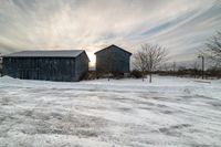 two barns are standing in the snow on a farm field at sunset and trees and buildings can be seen in the background