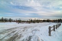 a snowy path by a fence with trees and clouds in the background and some cars parked on the snow