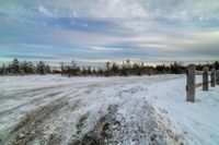 a snowy path by a fence with trees and clouds in the background and some cars parked on the snow