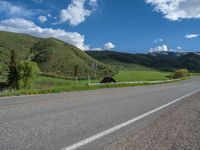 the empty road is going through a valley with mountains and grass in the background and two houses on both sides