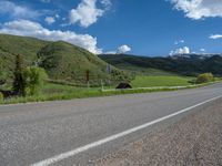 the empty road is going through a valley with mountains and grass in the background and two houses on both sides