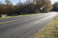 an empty rural country road passing a wooded area with fence posts in the distance and fall leaves on the ground