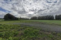 a field with an old building surrounded by trees and grass in the background, clouds are coming from behind