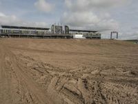 dirt track with some buildings and an event venue in the distance, in front of a field with a sky background