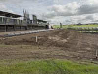 dirt track with some buildings and an event venue in the distance, in front of a field with a sky background