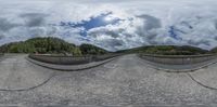 a 360 - view photo shows an empty road and mountains in the background, with trees, clouds and water in the foreground