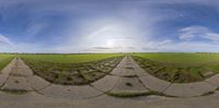 the 360 - lens view of a road with a grass field in the background and clouds overhead