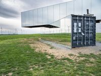 this container phone is upside down in the grass near wind turbines behind it with its reflection on the mirrored roof