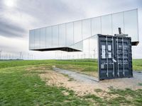 this container phone is upside down in the grass near wind turbines behind it with its reflection on the mirrored roof