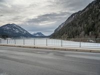 the mountains surrounding a lake are covered in snow as a man walks across it beside a fence