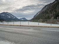 the mountains surrounding a lake are covered in snow as a man walks across it beside a fence