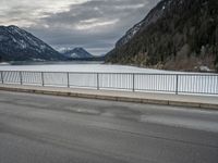 the mountains surrounding a lake are covered in snow as a man walks across it beside a fence