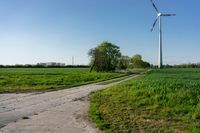 the wind turbine is on the edge of a dirt road in the fields with grass and some trees