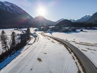 an aerial view of an alpine country with a winding road surrounded by trees and snow in the foreground, with sun reflecting off the mountain tops