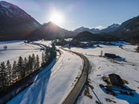 an aerial view of an alpine country with a winding road surrounded by trees and snow in the foreground, with sun reflecting off the mountain tops