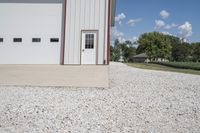 the side of a building with a motorcycle parked in front of it, in a gravel area in front of an open field