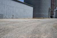 two large grain bins sitting next to a building with a dirt driveway and a metal structure