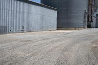 two large grain bins sitting next to a building with a dirt driveway and a metal structure