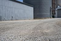 two large grain bins sitting next to a building with a dirt driveway and a metal structure