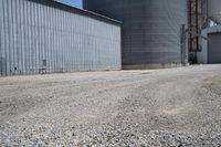 two large grain bins sitting next to a building with a dirt driveway and a metal structure