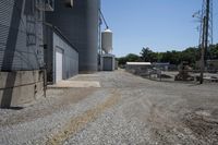 a gravel road in front of an industrial building and water tower with two silo on top