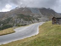 a small cabin sitting on top of a hill near a mountain road going past a tall mountain