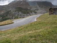 a small cabin sitting on top of a hill near a mountain road going past a tall mountain