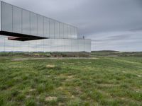 a mirrored structure in the middle of a grassy field overlooking a river with buildings in the background