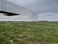 a mirrored structure in the middle of a grassy field overlooking a river with buildings in the background